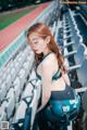 A woman in a sports bra top and leggings sitting in a stadium.